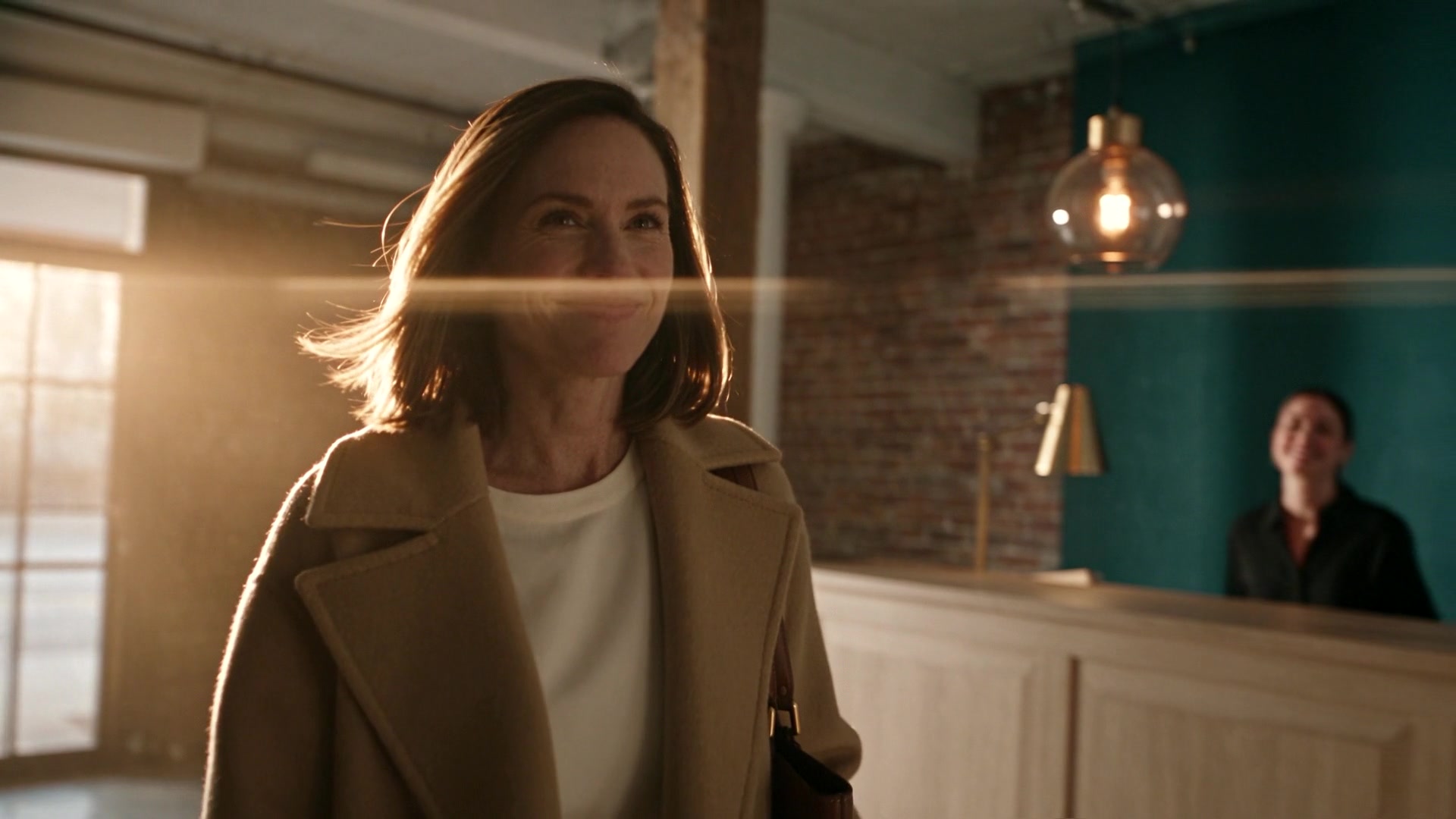 A smiling woman in a camel coat arriving at a reception desk in warm light